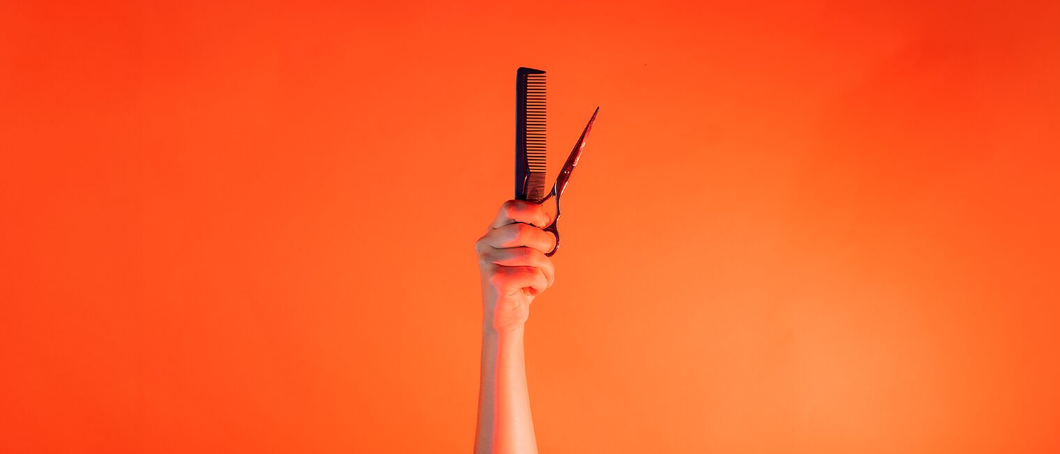 Ein Arm ist vor orangenem Hintergrund senkrecht nach oben gestreckt und hält in der Hand einen Kamm und eine Schere. 