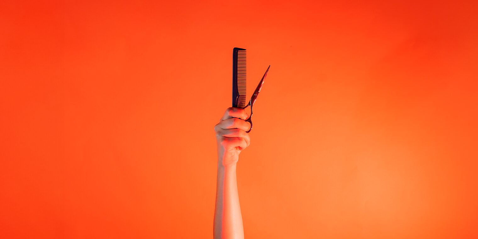 Ein Arm ist vor orangenem Hintergrund senkrecht nach oben gestreckt und hält in der Hand einen Kamm und eine Schere. 