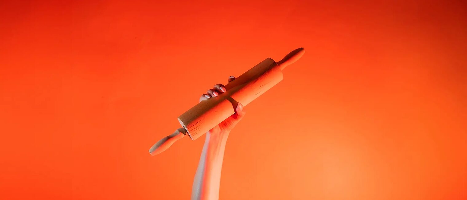 Ein Arm ist vor orangenem Hintergrund senkrecht nach oben gestreckt und in der Hand wird ein Nudelholz gehalten.