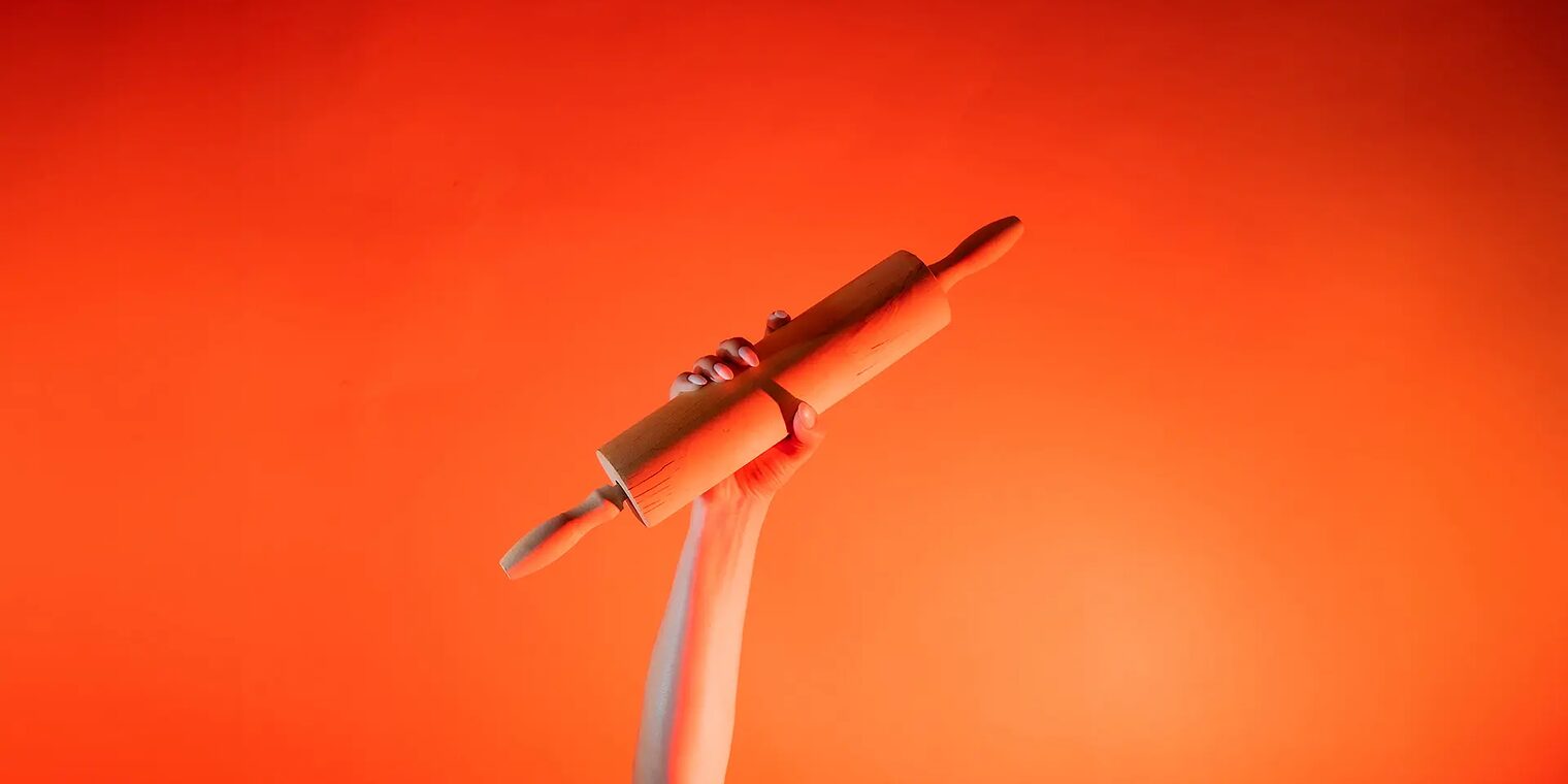 Ein Arm ist vor orangenem Hintergrund senkrecht nach oben gestreckt und in der Hand wird ein Nudelholz gehalten.
