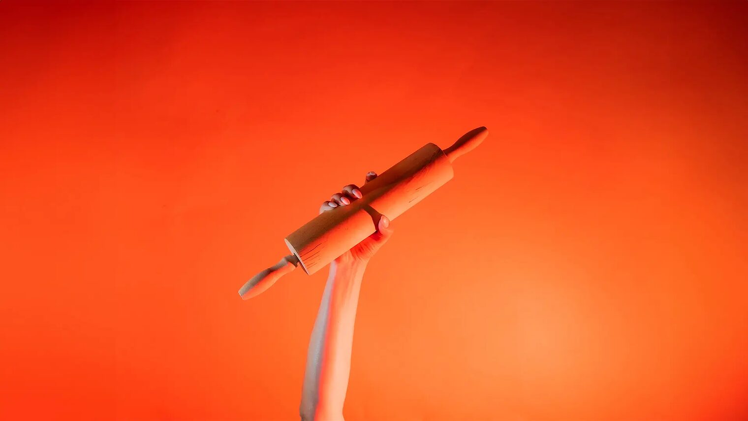 Ein Arm ist vor orangenem Hintergrund senkrecht nach oben gestreckt und in der Hand wird ein Nudelholz gehalten.