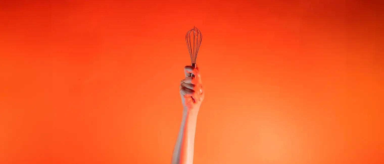Ein Arm ist vor orangenem Hintergrund senkrecht nach oben gestreckt und in der Hand wird ein Schneebesen gehalten.