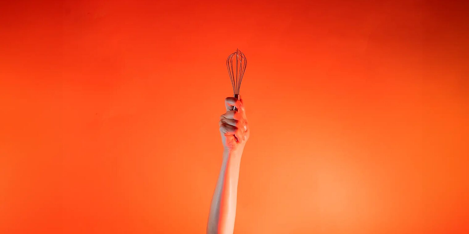 Ein Arm ist vor orangenem Hintergrund senkrecht nach oben gestreckt und in der Hand wird ein Schneebesen gehalten.