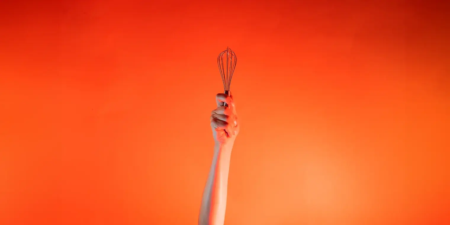 Ein Arm ist vor orangenem Hintergrund senkrecht nach oben gestreckt und in der Hand wird ein Schneebesen gehalten.
