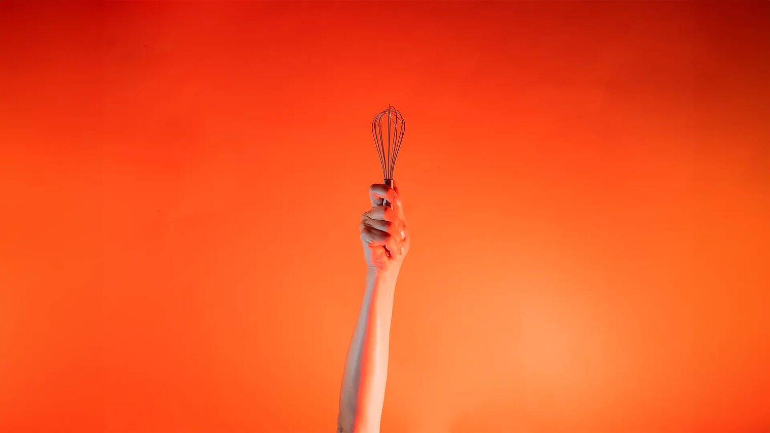 Ein Arm ist vor orangenem Hintergrund senkrecht nach oben gestreckt und in der Hand wird ein Schneebesen gehalten.