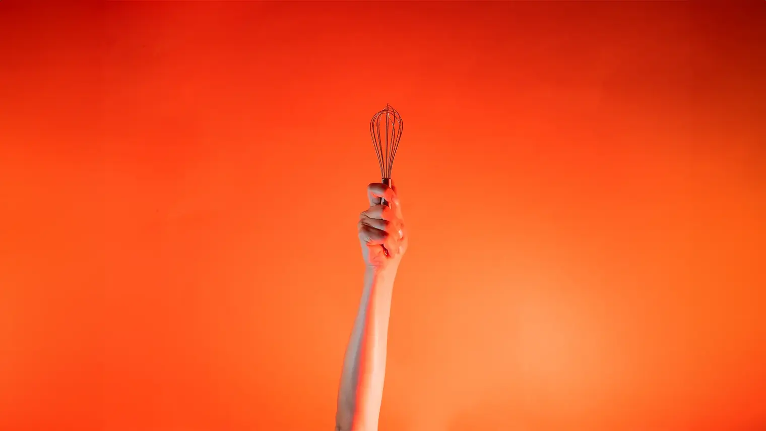 Ein Arm ist vor orangenem Hintergrund senkrecht nach oben gestreckt und in der Hand wird ein Schneebesen gehalten.