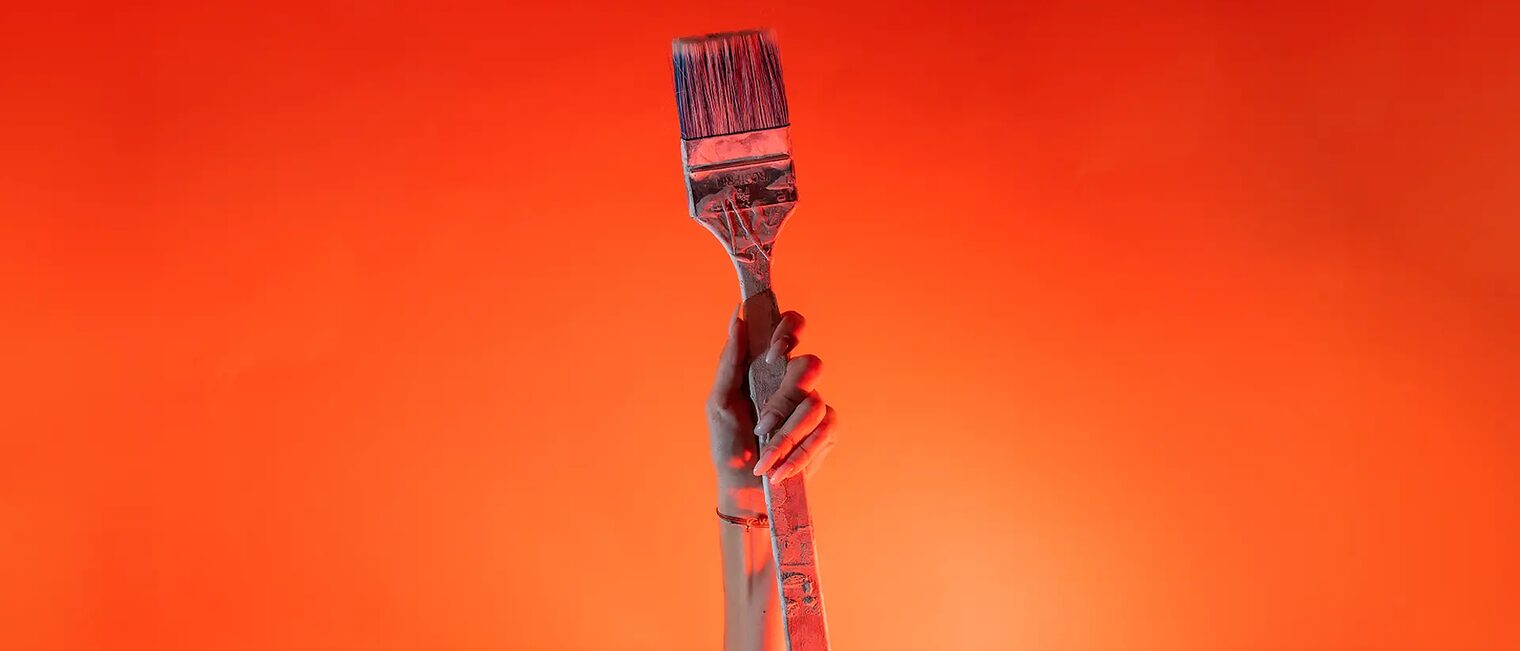 Ein Arm ist vor orangenem Hintergrund senkrecht nach oben gestreckt und in der Hand wird ein Malerpinsel gehalten.