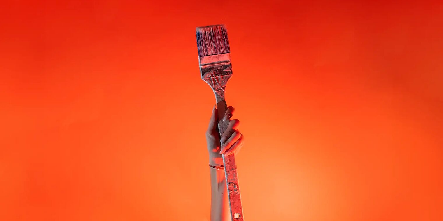 Ein Arm ist vor orangenem Hintergrund senkrecht nach oben gestreckt und in der Hand wird ein Malerpinsel gehalten.
