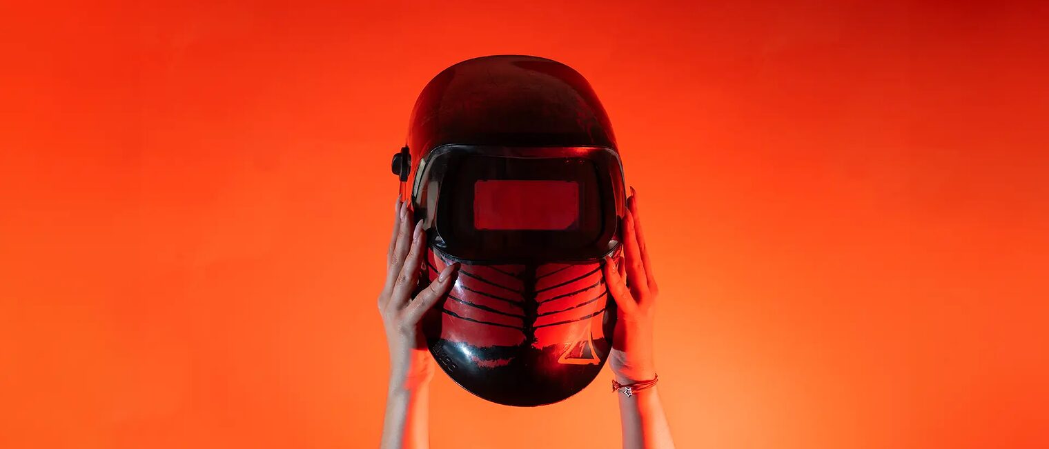 Zwei Hände halten vor orangenem Hintergrund einen Schweißhelm senkrecht nach oben.