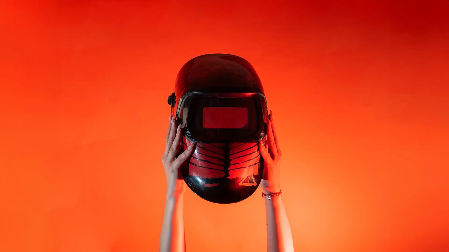 Zwei Hände halten vor orangenem Hintergrund einen Schweißhelm senkrecht nach oben.