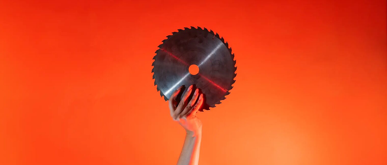 Ein Arm ist vor orangenem Hintergrund senkrecht nach oben gestreckt und in der Hand wird ein rundes Sägeblatt gehalten.