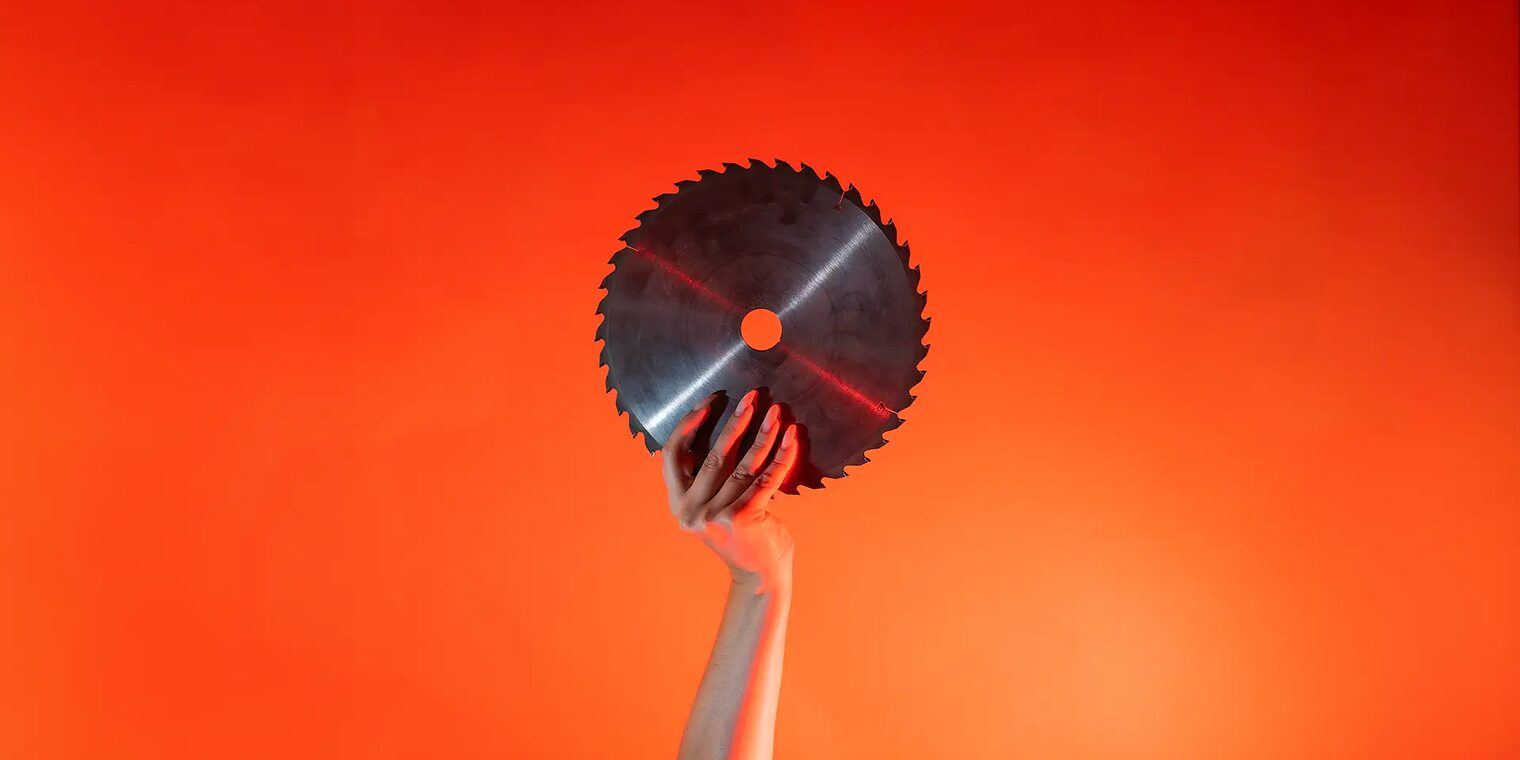 Ein Arm ist vor orangenem Hintergrund senkrecht nach oben gestreckt und in der Hand wird ein rundes Sägeblatt gehalten.