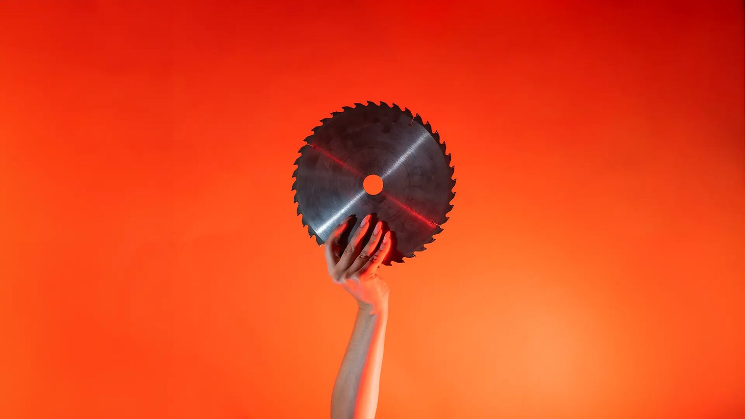 Ein Arm ist vor orangenem Hintergrund senkrecht nach oben gestreckt und in der Hand wird ein rundes Sägeblatt gehalten.