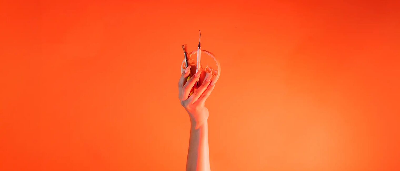 Eine Hand ist vor orangenem Hintergrund senkrecht nach oben gestreckt und hält: Wachspinsel, LeCron-Werkzeug, 3D-Druckzahn.