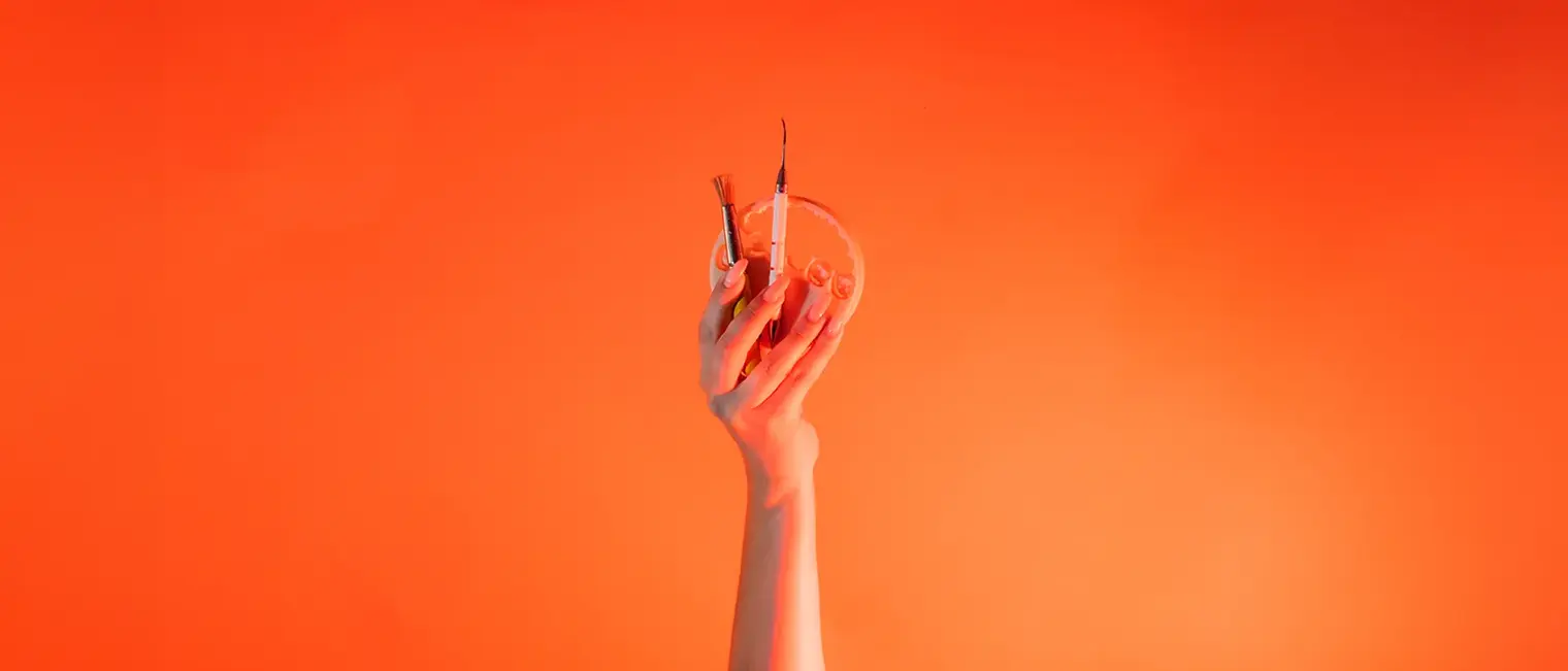 Eine Hand ist vor orangenem Hintergrund senkrecht nach oben gestreckt und hält: Wachspinsel, LeCron-Werkzeug, 3D-Druckzahn.