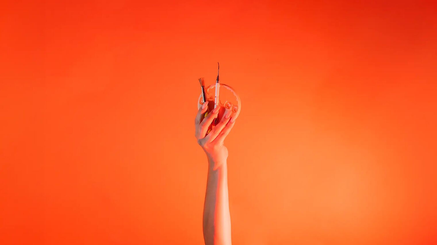 Eine Hand ist vor orangenem Hintergrund senkrecht nach oben gestreckt und hält: Wachspinsel, LeCron-Werkzeug, 3D-Druckzahn.