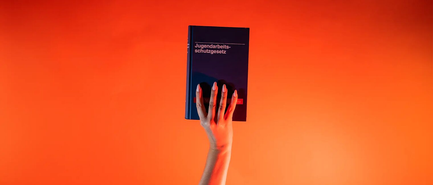 Ein Arm ist vor orangenem Hintergrund senkrecht nach oben gestreckt und hält in der Hand das Jugendarbeitsschutzgesetz.