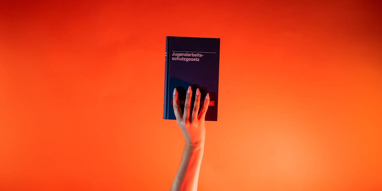 Ein Arm ist vor orangenem Hintergrund senkrecht nach oben gestreckt und hält in der Hand das Jugendarbeitsschutzgesetz.