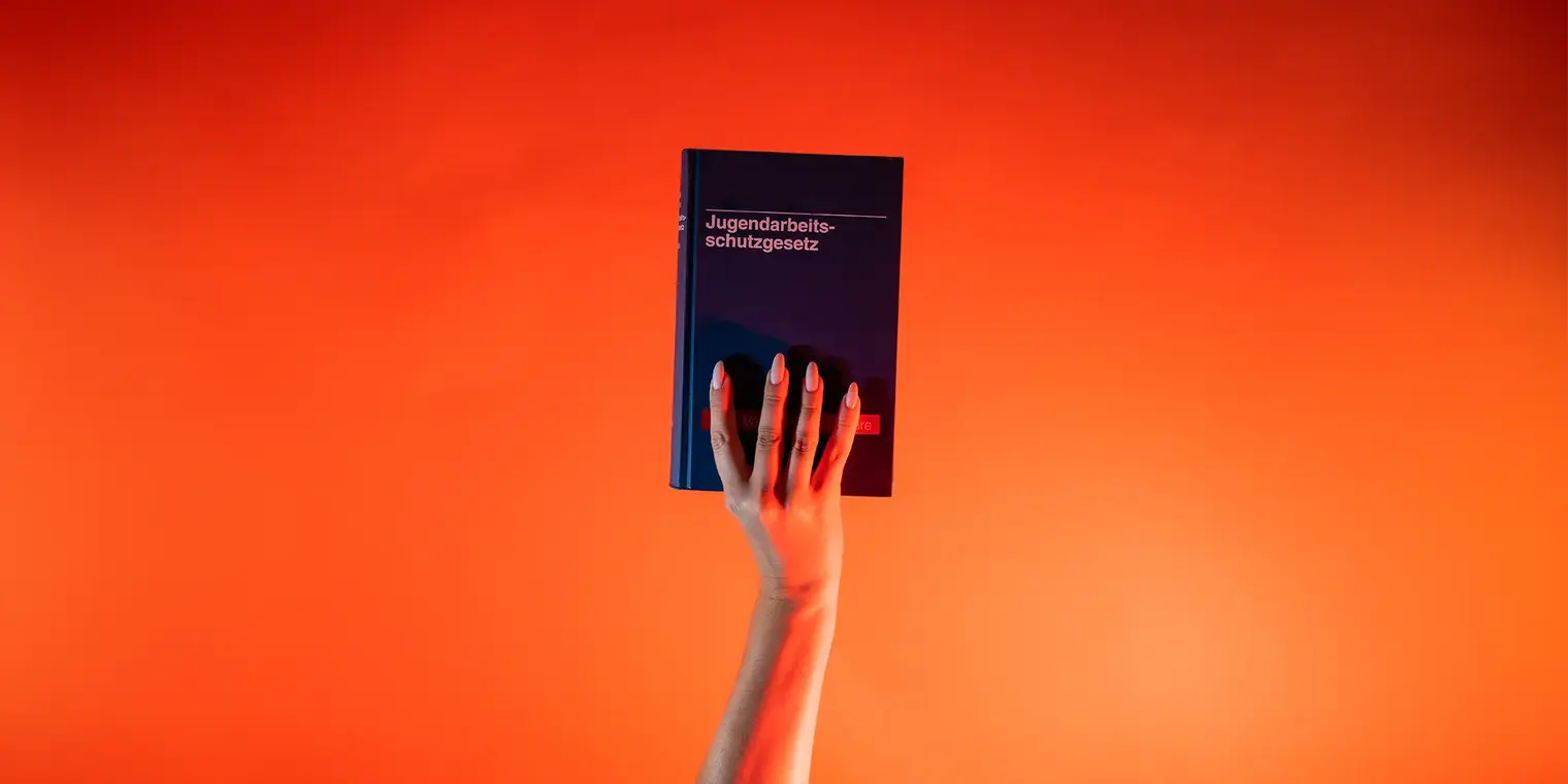 Ein Arm ist vor orangenem Hintergrund senkrecht nach oben gestreckt und hält in der Hand das Jugendarbeitsschutzgesetz.