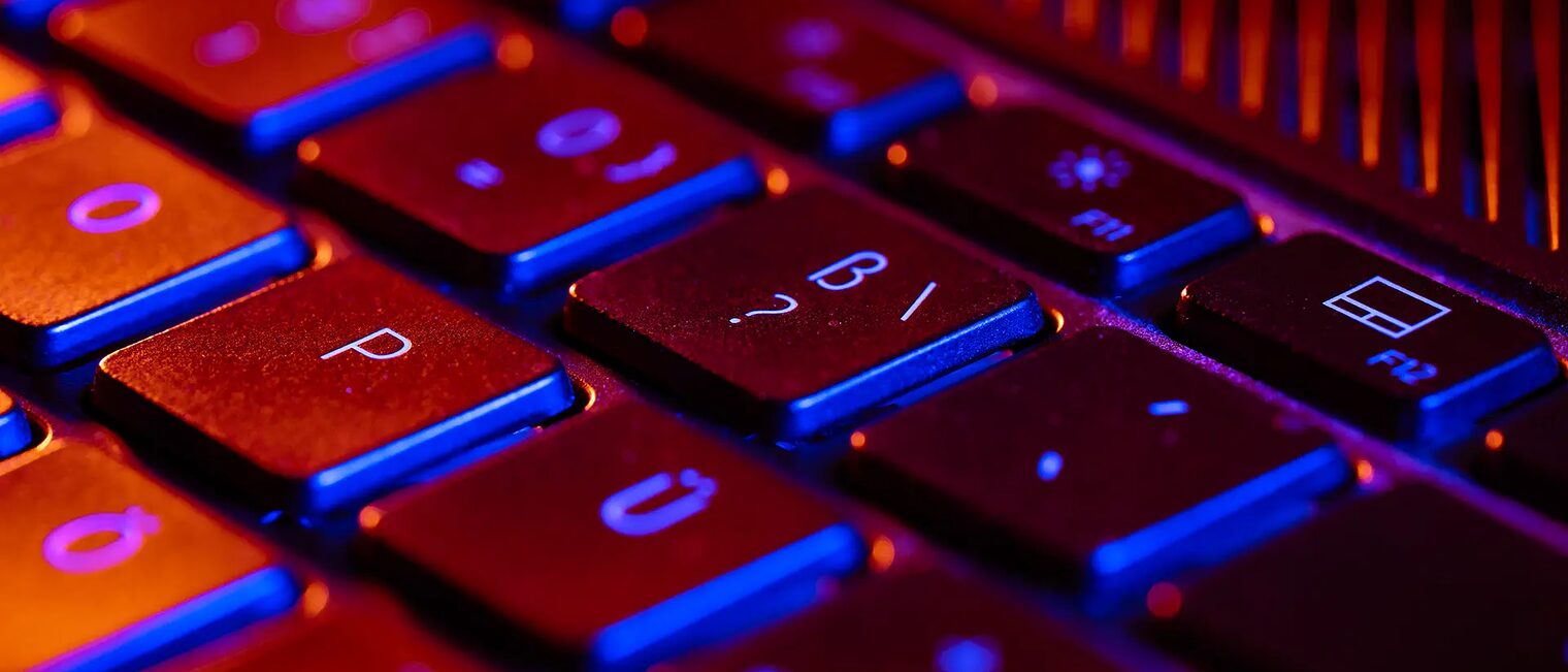 Die Tastatur eines Laptops wird orange und blau angeleuchtet. Im Zentrum und Focus des Fotos: Die Fragezeichentaste, als Symbol für die FAQs.