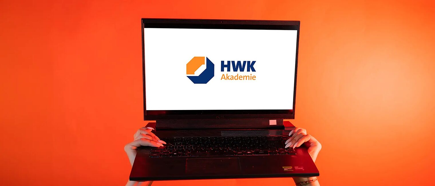 Ein Foto mit dem Akademie-Marken-Logo für die Seite "über uns". Zwei Hände heben einen Laptop vor orangenem Hintergrund in die Luft. Der Bildschirm ist weiß und mittig liegt das Logo Akademie.