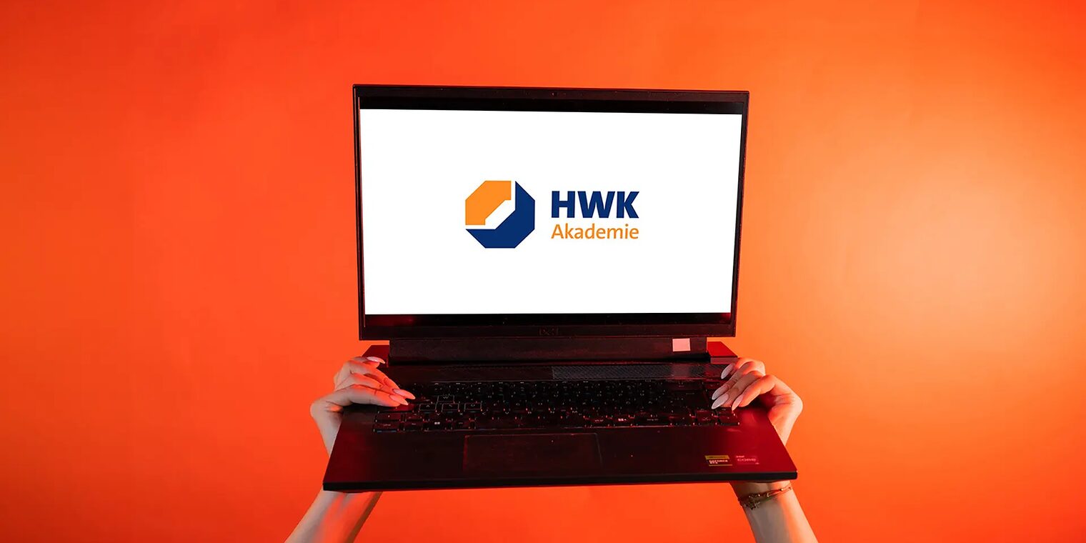Zwei Hände heben einen Laptop vor orangenem Hintergrund in die Luft. Der Bildschirm ist weiß und mittig liegt das Logo Akademie.