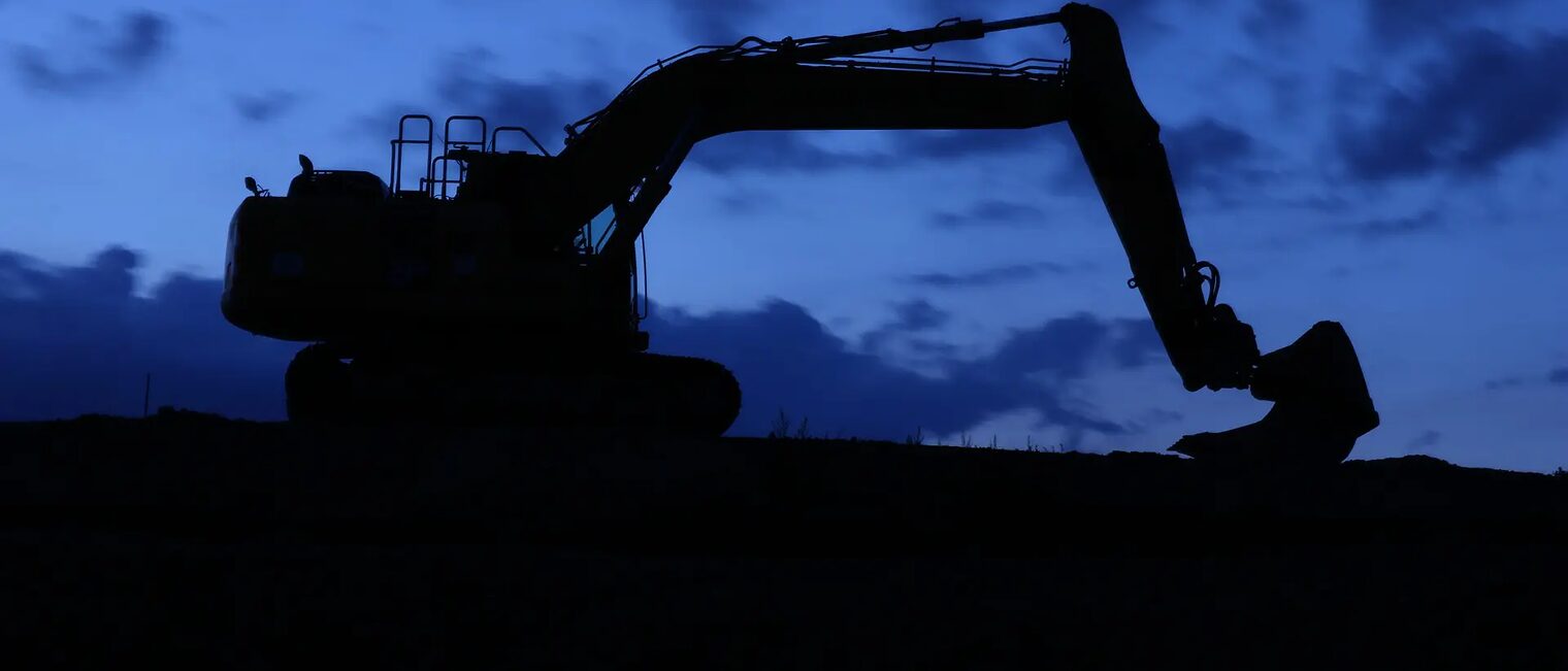 Foto: Info zur Baustelle Foto eines Baggers in der Nacht. Im Hintergrund der dunkelblaue Abendhimmel. Im Vordergrund die Silhouette des Baggers auf einer Straße.