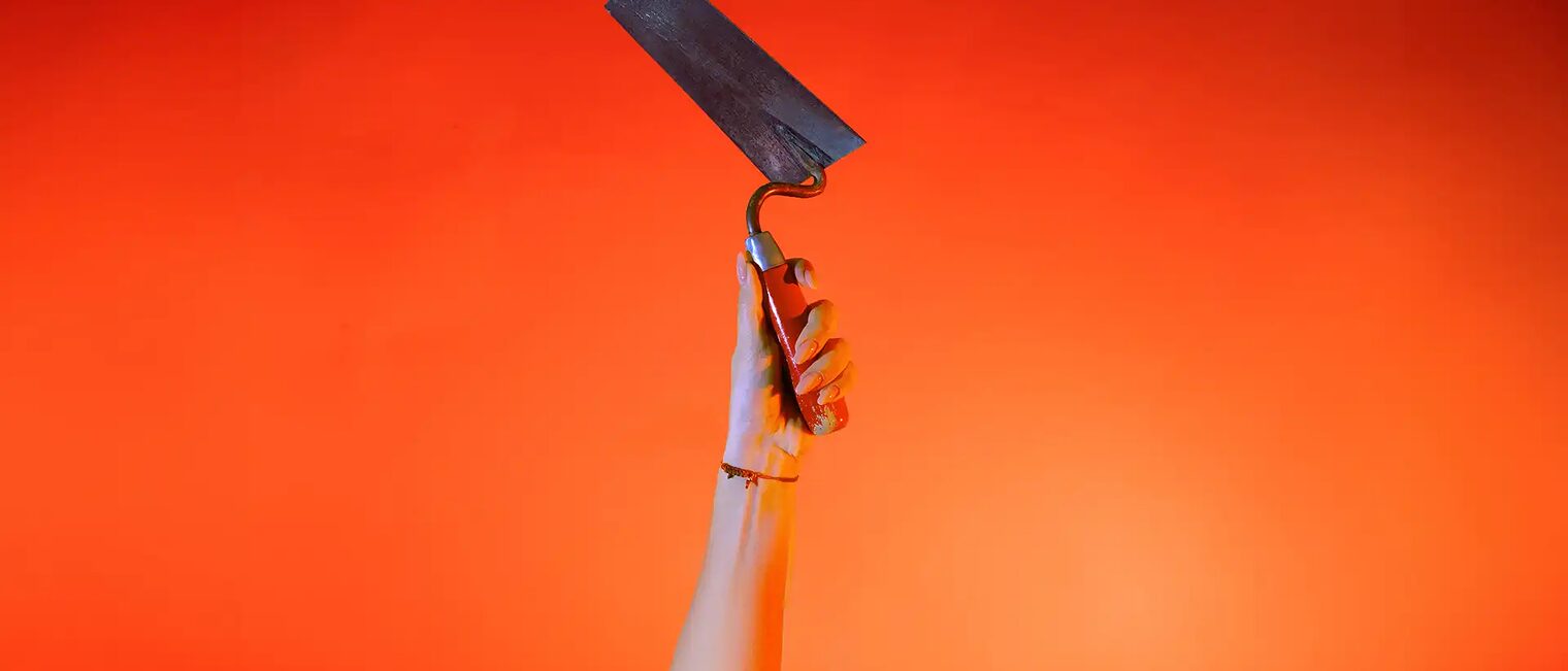 Ein Hand streckt vor orangenem Hintergrund eine Maurerkelle senkrecht nach oben.