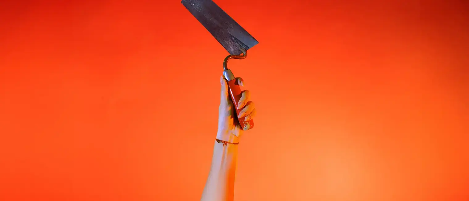 Ein Hand streckt vor orangenem Hintergrund eine Maurerkelle senkrecht nach oben.