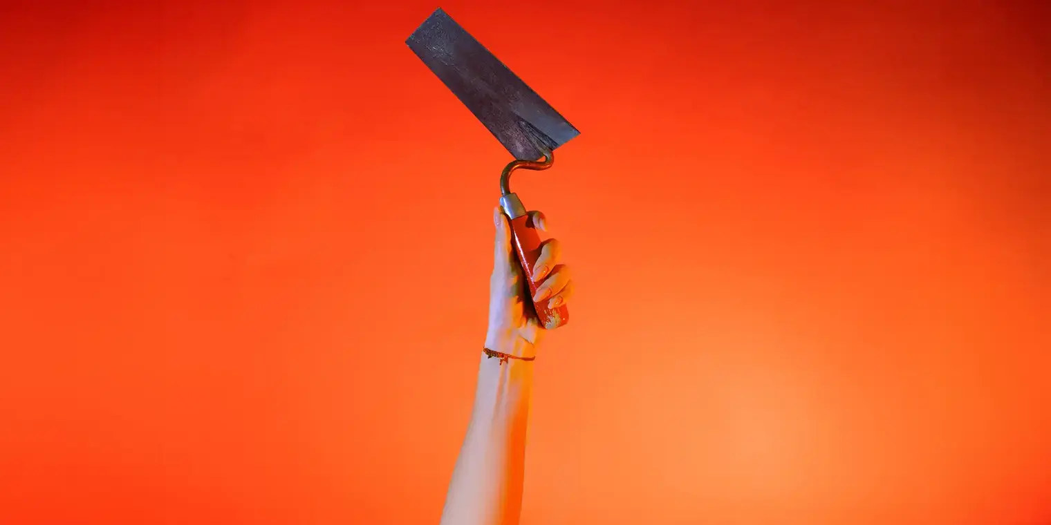Ein Hand streckt vor orangenem Hintergrund eine Maurerkelle senkrecht nach oben.
