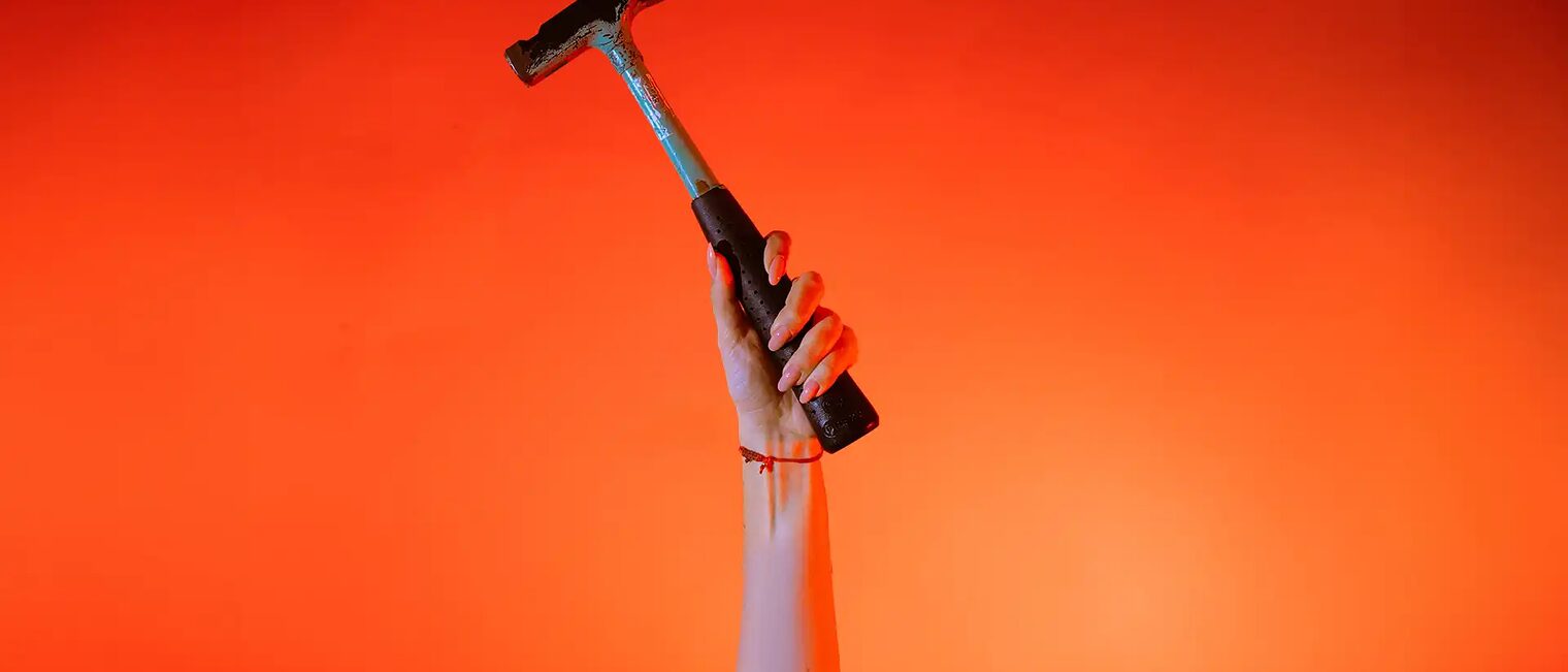 Eine Hand hält einen Zimmererhammer vor orangenem Hintergrund senkrecht in die Höhe.