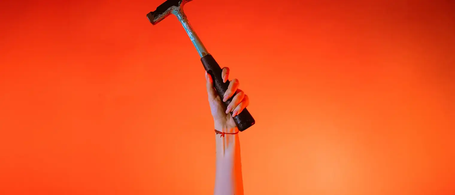 Eine Hand hält einen Zimmererhammer vor orangenem Hintergrund senkrecht in die Höhe.