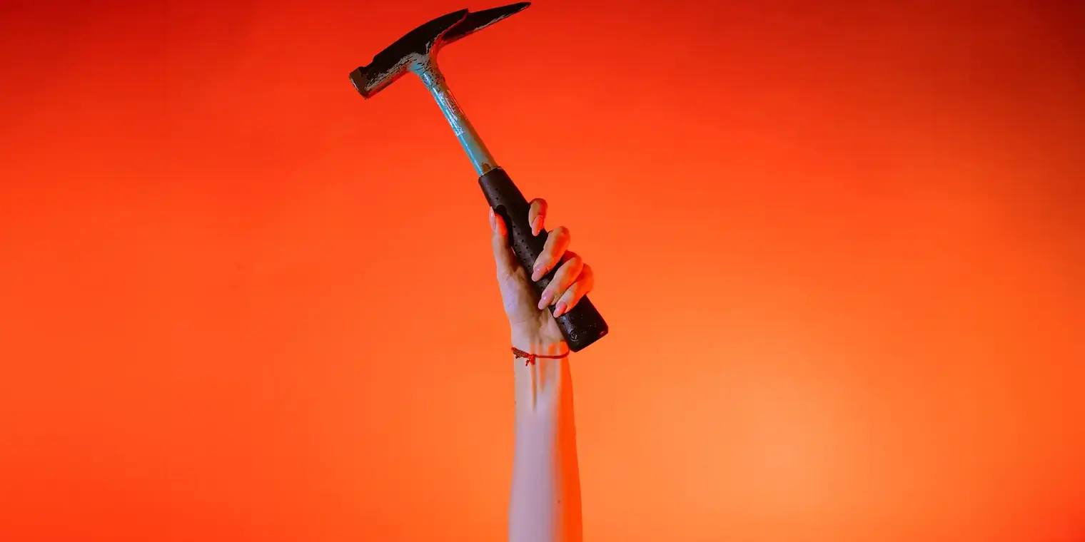 Eine Hand hält einen Zimmererhammer vor orangenem Hintergrund senkrecht in die Höhe.