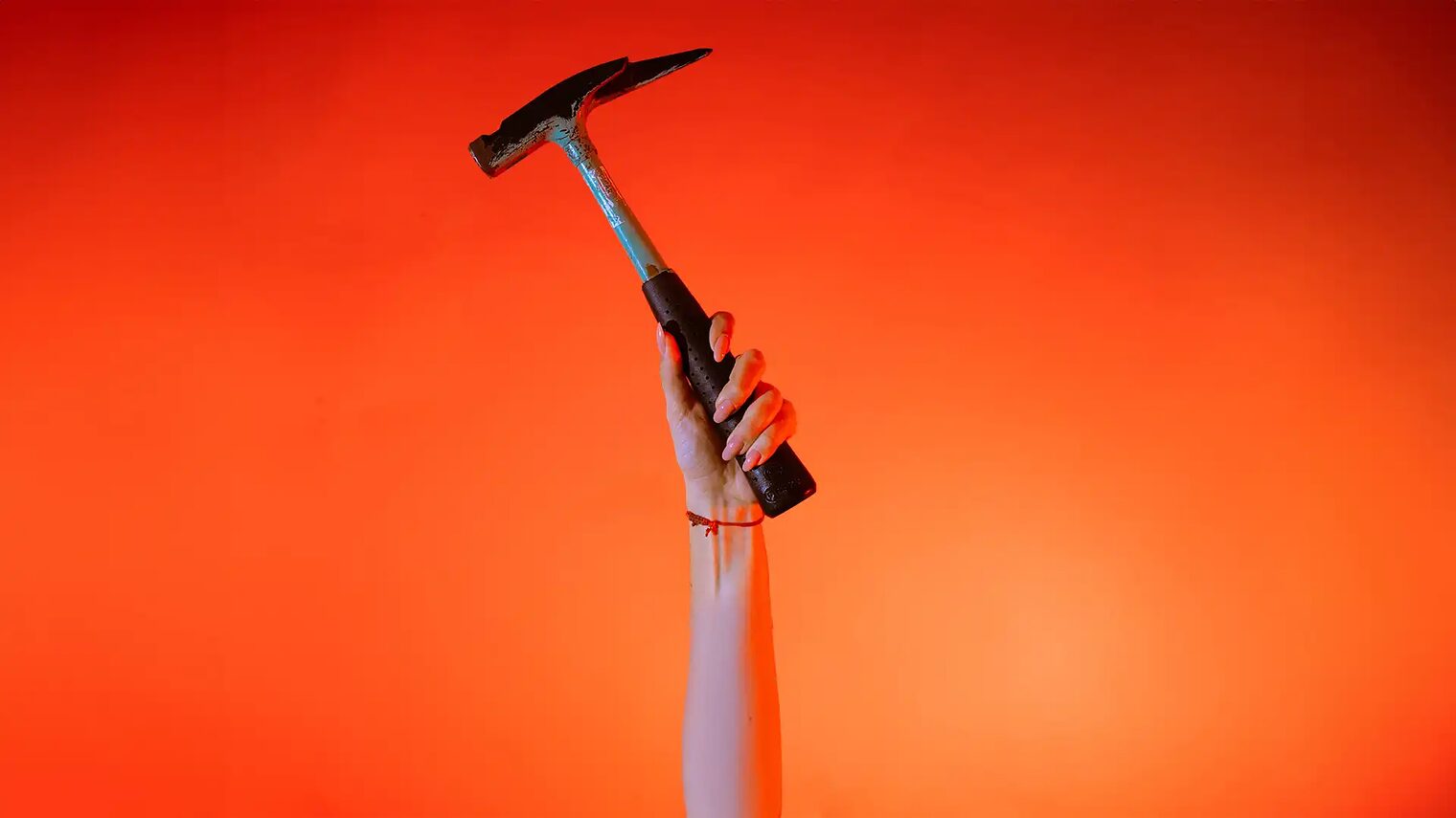 Eine Hand hält einen Zimmererhammer vor orangenem Hintergrund senkrecht in die Höhe.