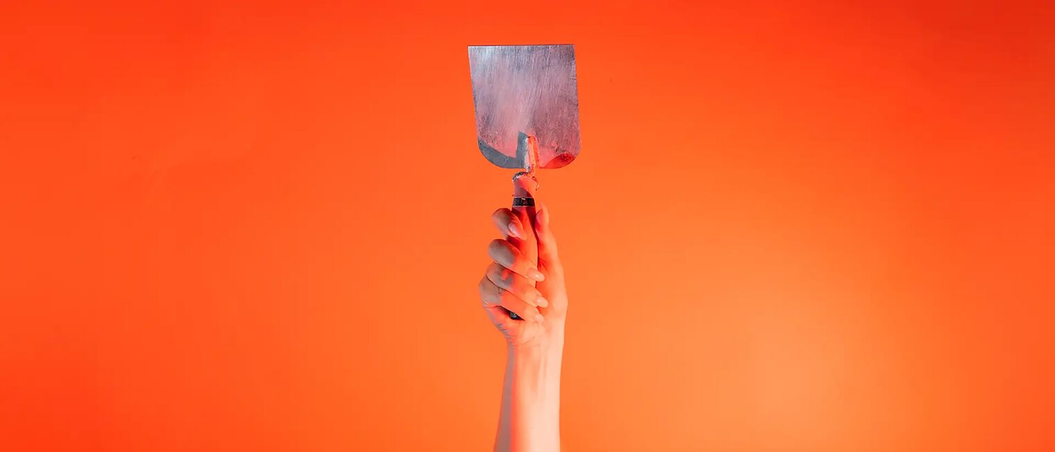 Eine Hand steckt vor orangenem Hintergrund eine kleine Stuckateur Kelle senkrecht nach oben.
