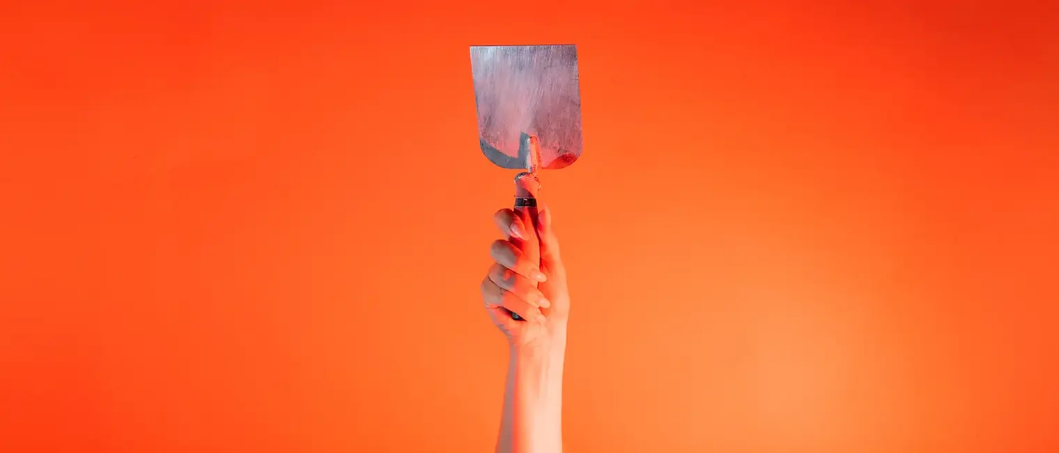 Eine Hand steckt vor orangenem Hintergrund eine kleine Stuckateur Kelle senkrecht nach oben.