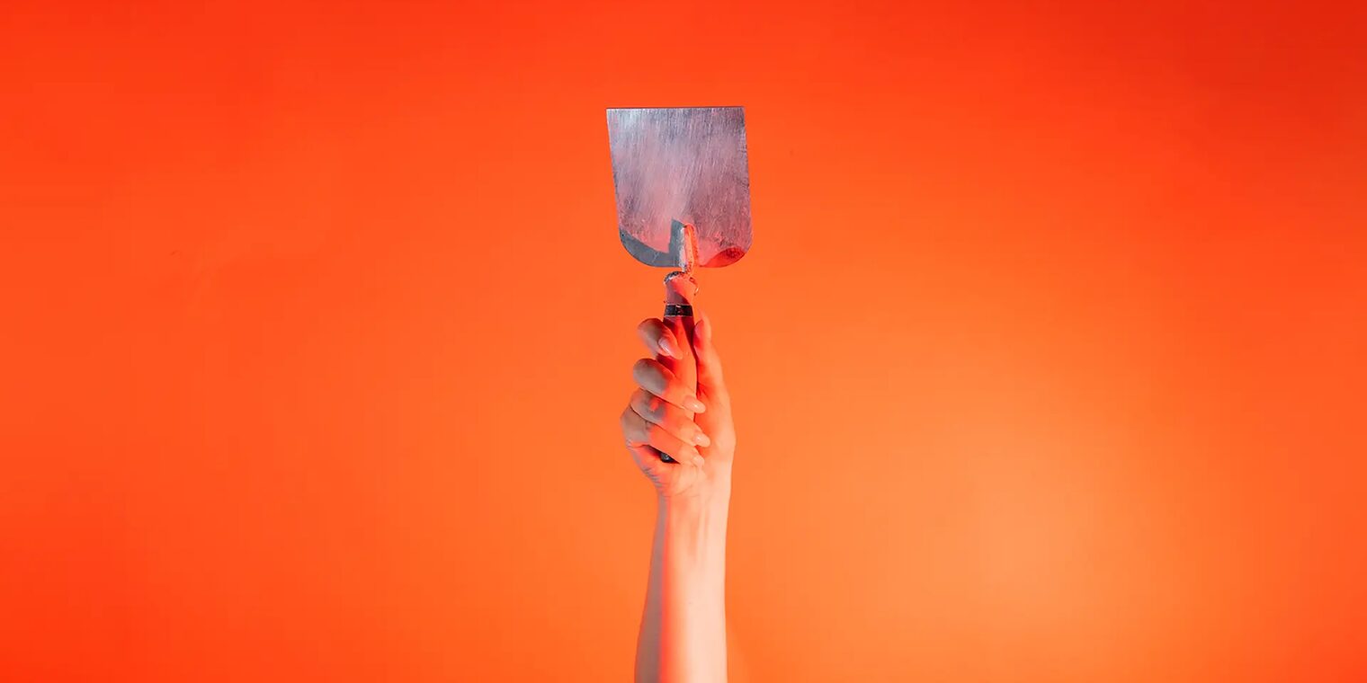 Eine Hand steckt vor orangenem Hintergrund eine kleine Stuckateur Kelle senkrecht nach oben.