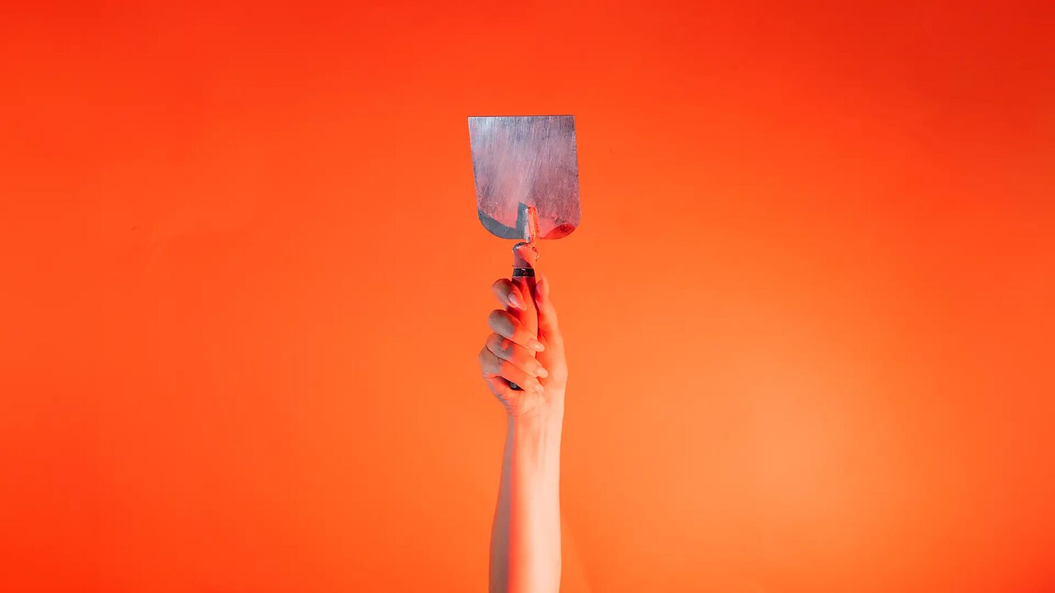 Eine Hand steckt vor orangenem Hintergrund eine kleine Stuckateur Kelle senkrecht nach oben.