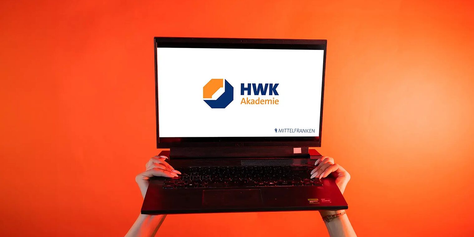 Zwei Hände heben einen Laptop in die Luft. Der Bildschirm ist weiß und mittig liegt das Logo Akademie.